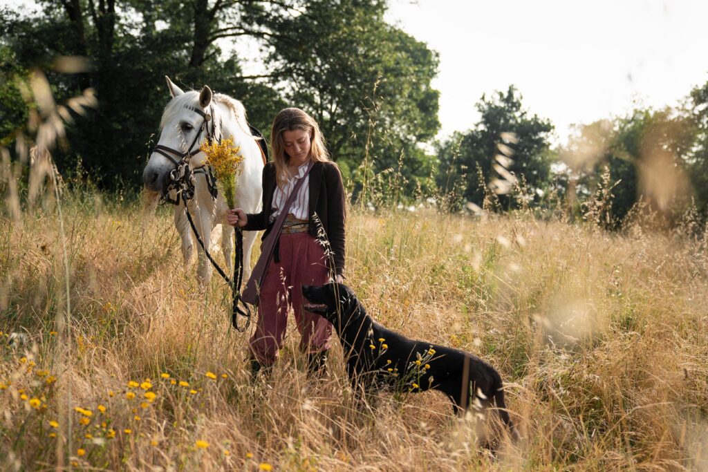 Woman standing in a sunny field holding a grey horse and a yellow bunch of flowers next to a black labrador dog